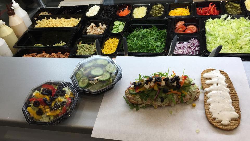 Frisklavet sandwich med grøntsager og salat, samt to salater i plastikbeholdere, på en køkkenbænk.