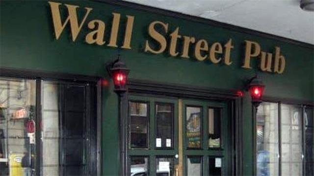 Indgangen til Wall Street Pub med grøn facade og gyldne bogstaver. Røde lamper lyser op ved døren.
