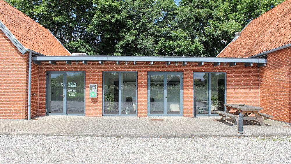 En rød murstensbygning med store glasdøre og et picnicbord foran. Grønne træer i baggrunden.