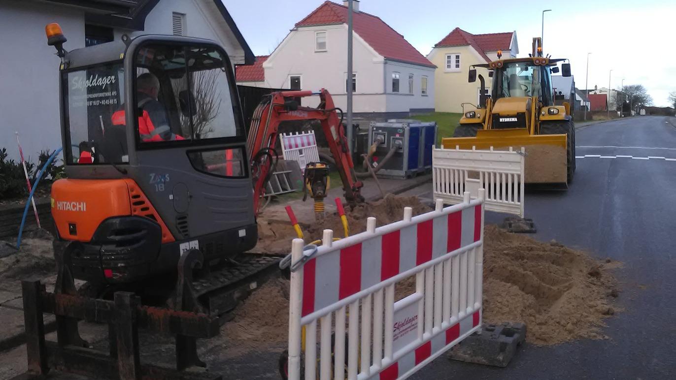 Gravemaskine og bulldozer arbejder på en vej, omgivet af afspærringshegn og bygninger i baggrunden.