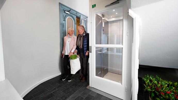 To personer, en mand og en kvinde, står ved en elevator i en moderne gang med planter i baggrunden.