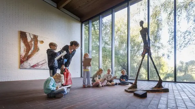 Børn sidder på gulvet i et galleri og ser på en skulptur, mens en voksen viser dem noget på en tablet.