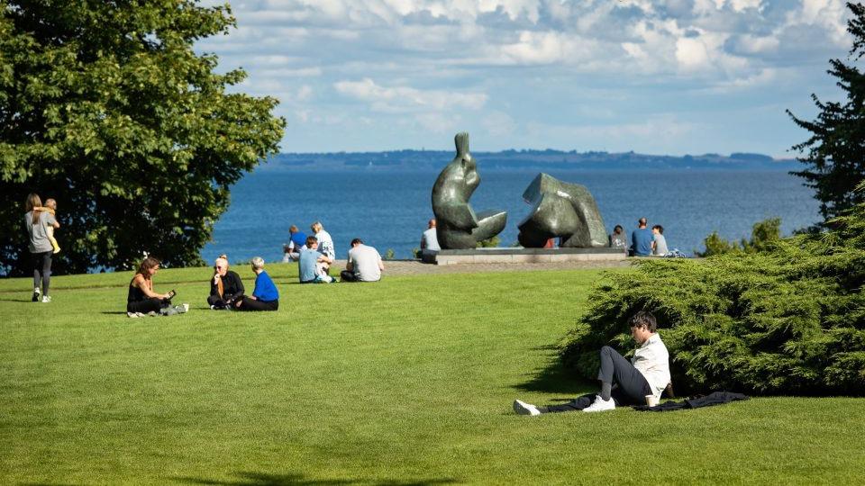 Mennesker sidder på græsset i en park med skulpturer og udsigt over vandet og himlen.
