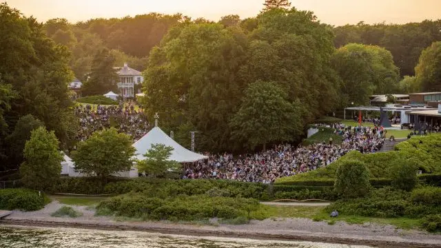 En stor folkemængde samles ved en udendørs koncert i en park med grønne træer og en hvid telt.