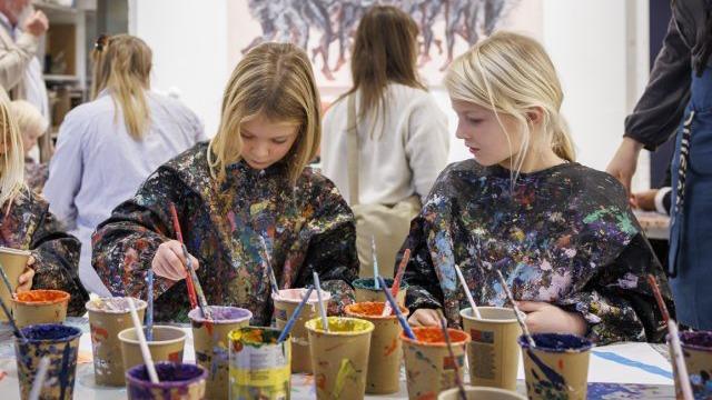 To piger maler med farver i papkrus under en kreativ workshop. De bærer malerkapper og koncentrerer sig om deres kunst.