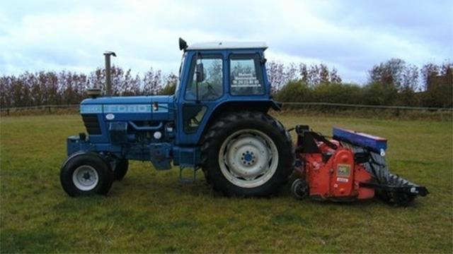 Blå traktor med rød fræser på en græsmark under en skyet himmel.