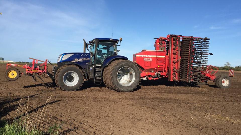 En blå traktor med store dæk trækker en rød såmaskine over en mark under en klar blå himmel.