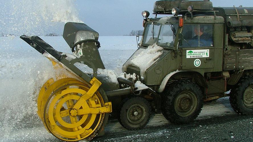 En sneslynge rydder sne fra vejen med kraftfulde sneskyder og gule skiver i et vinterlandskab.