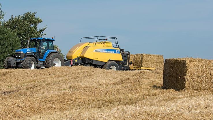 Traktor og høstmaskine arbejder på en mark med halmballer under en blå himmel.