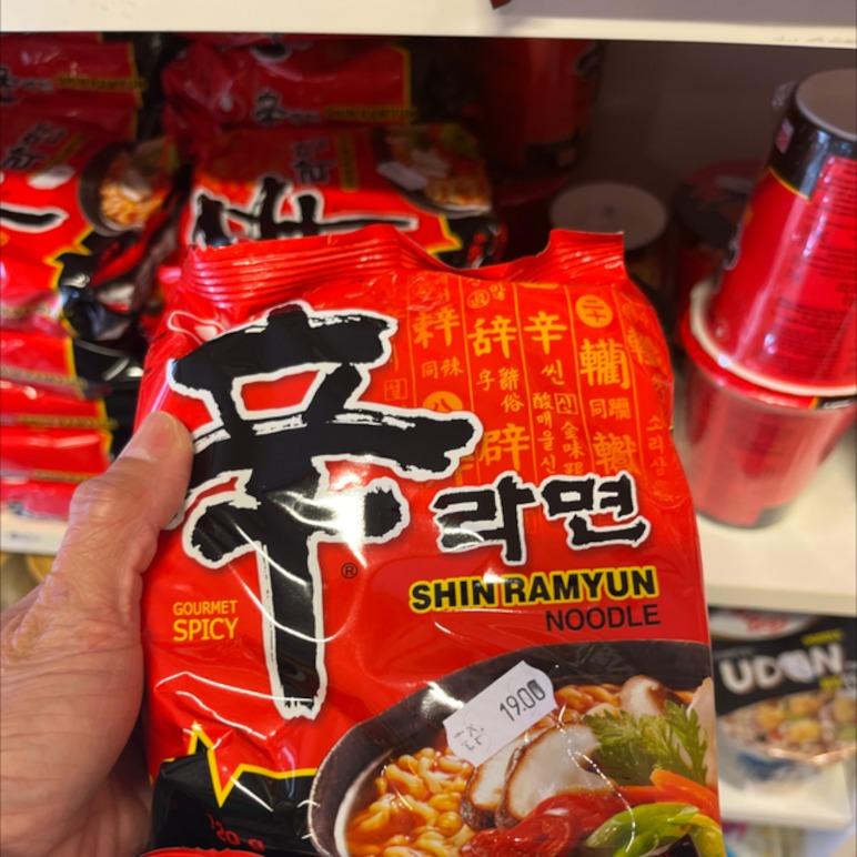 En person holder en pakke Shin Ramyun nudler med rød emballage og krydret tekst på hylden i en butik.