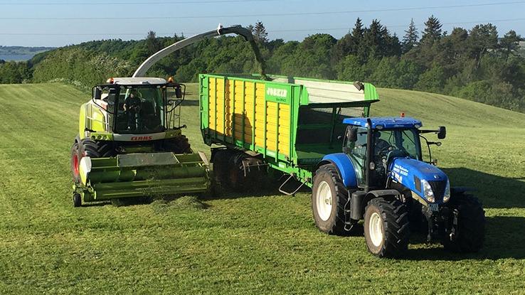 En traktor og en høstmaskine arbejder på en grøn mark, mens de høster græs til foder.