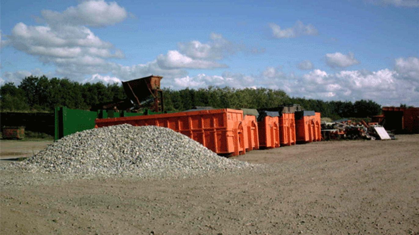 Affaldscontainere i orange farve står på en grusplads under en blå himmel med skyer.