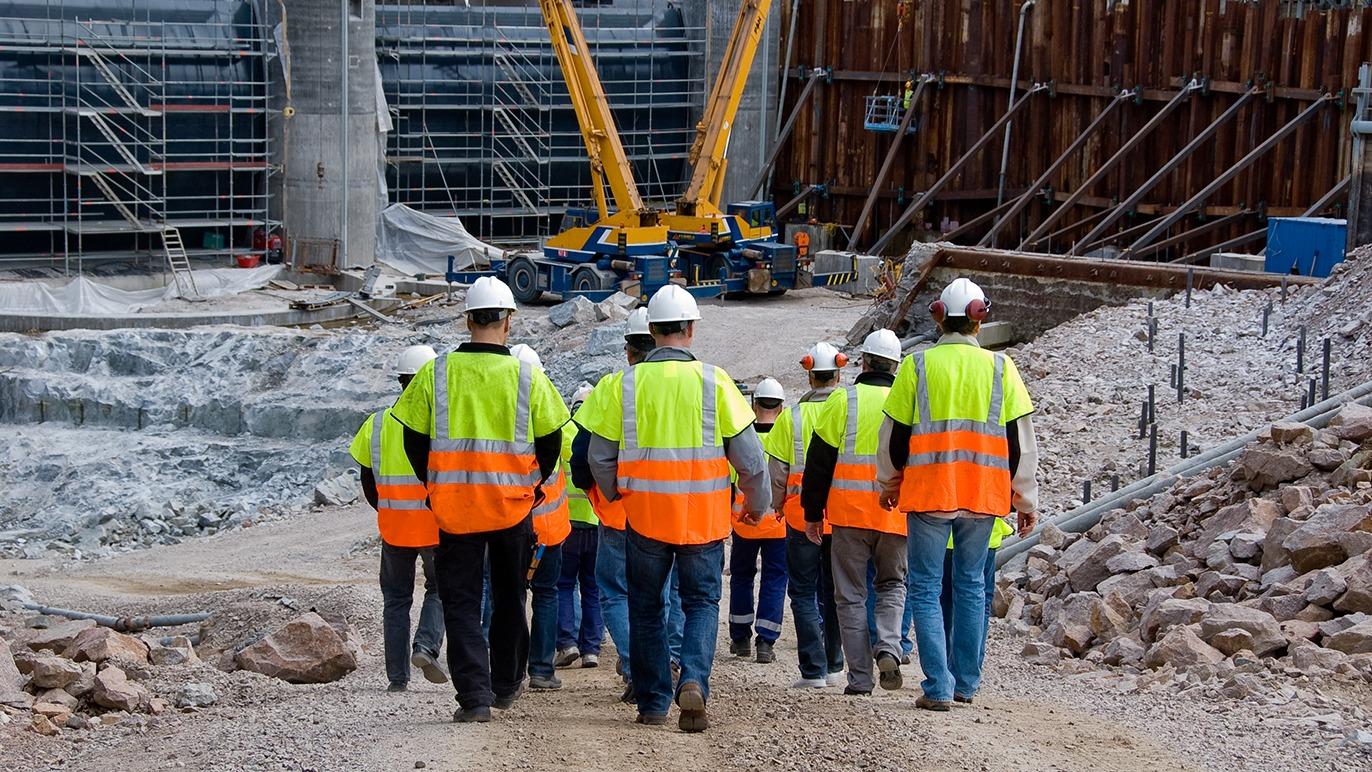Gruppe af arbejdere iført sikkerhedshjelme og refleksveste på en byggeplads med kraner og konstruktion.