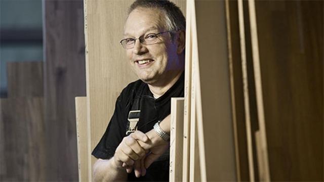 En smilende håndværker står mellem træplader i et værksted, iført arbejdsbukser og briller.