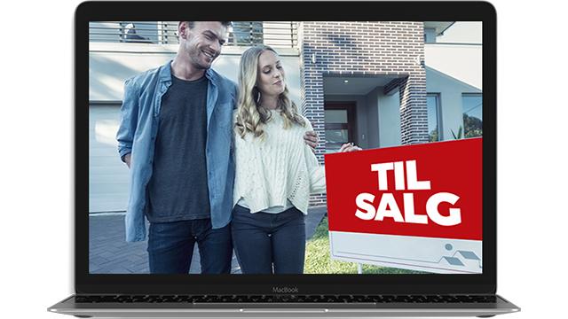 Et par står foran et hus med en "TIL SALG" skilt i hånden, mens de smiler til kameraet.