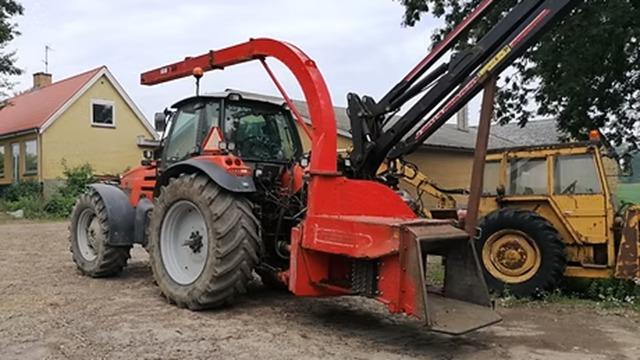 Rød traktor med kran og skovl parkeret ved en gård med bygninger i baggrunden.