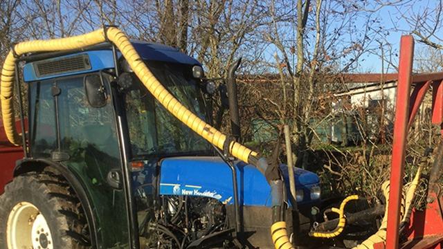 Traktor med gule slanger tilkoblet, parkeret i en have med træer og bygninger i baggrunden.