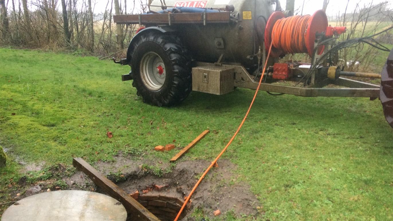 Traktor med slange ved en brønd, hvor der er gravet et hul i jorden, omgivet af græs og træer.