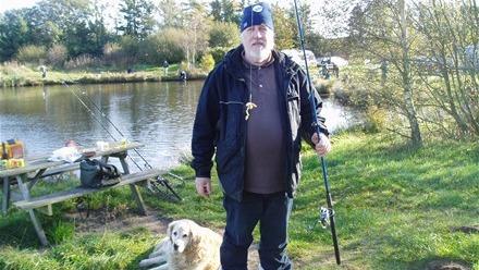 En mand står ved en sø med en fiskestang, mens en golden retriever ligger ved hans fødder.