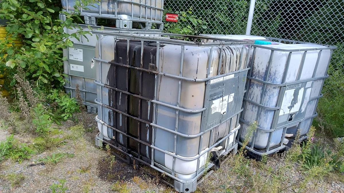 Stabel af plasttanke til opbevaring af væsker, placeret ved et hegn i et grønt område.