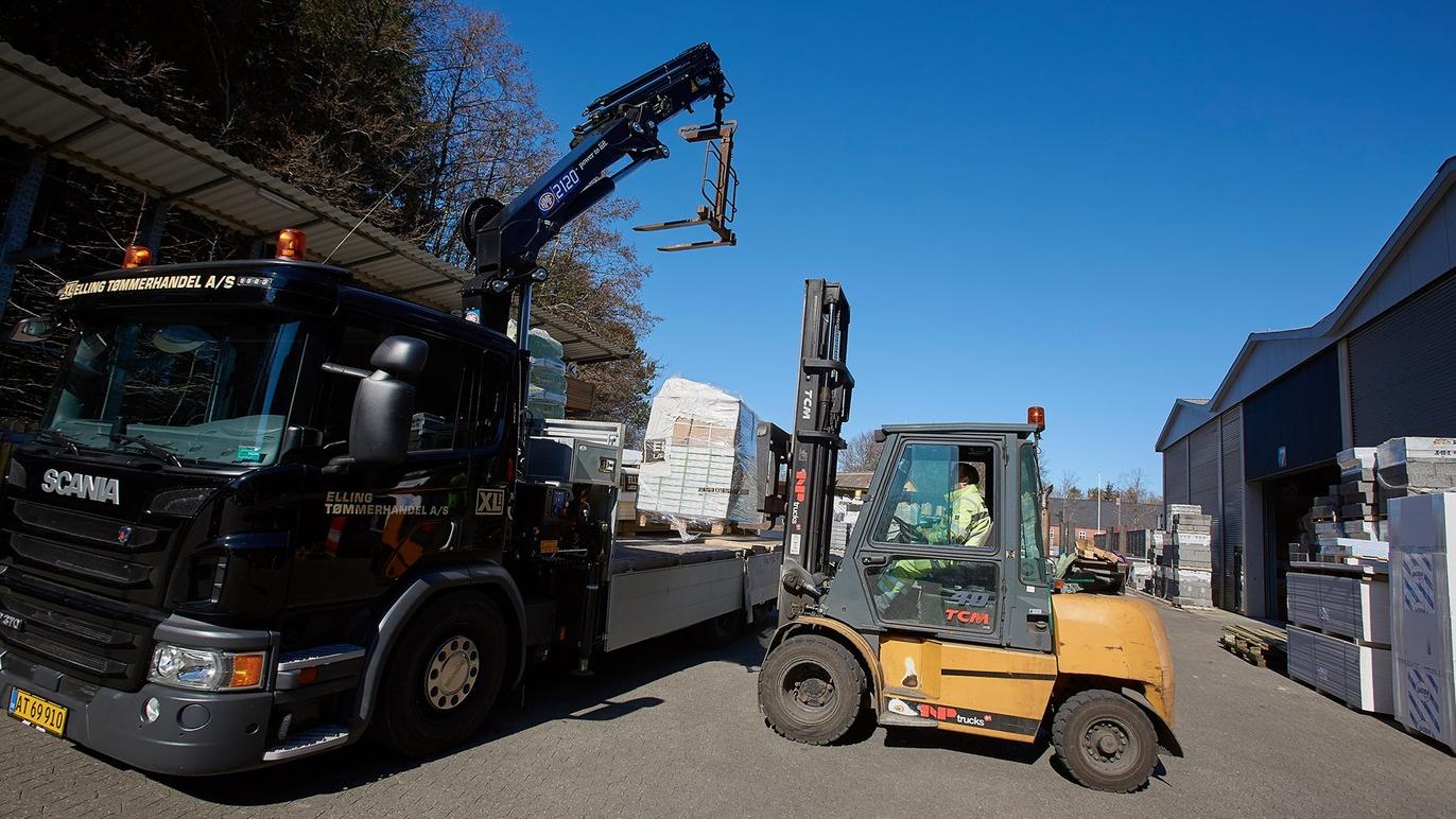 Lastbil og gaffeltruck arbejder sammen i en byggeplads under en klar blå himmel.