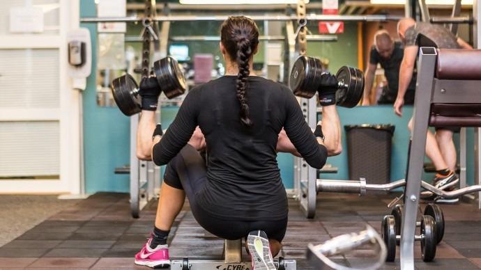 Kvinde træner med håndvægte i fitnesscenter, mens andre personer træner i baggrunden.