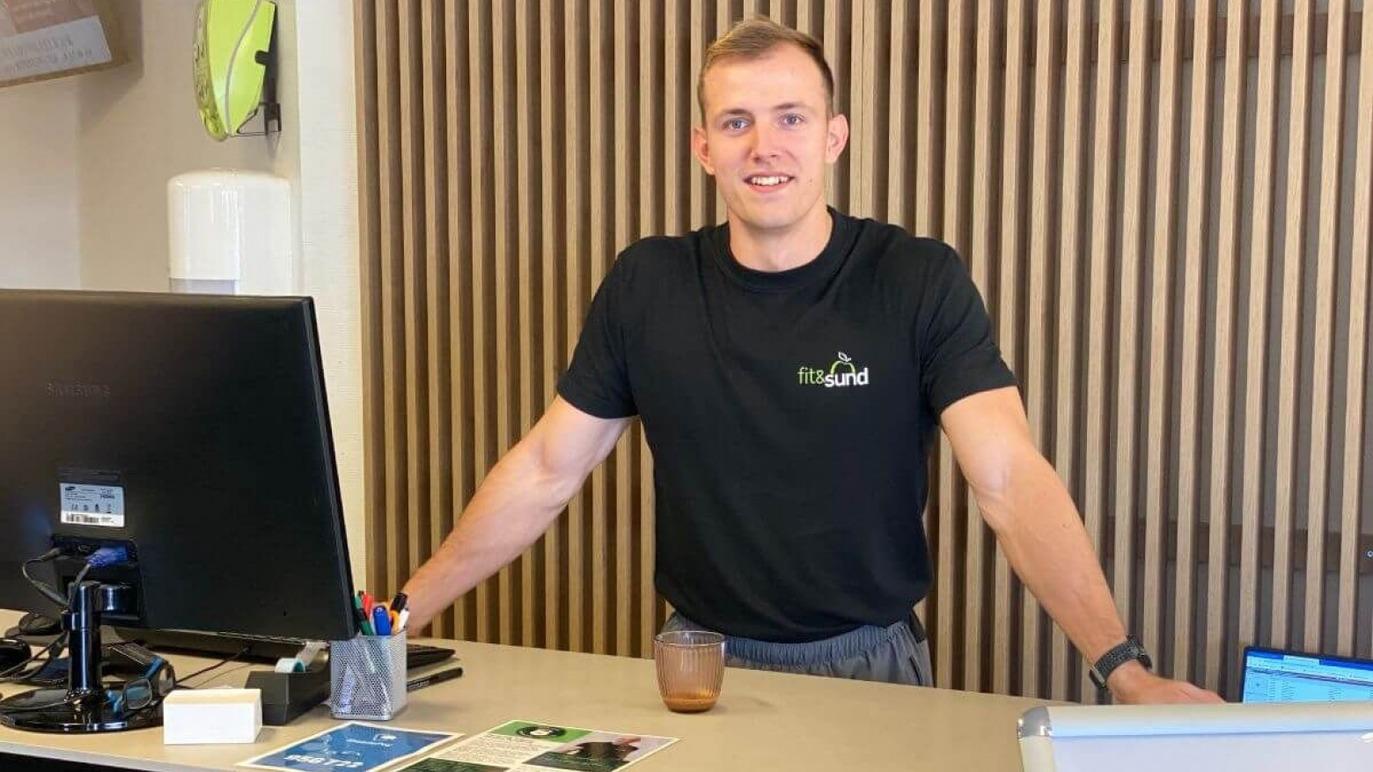 En ung mand står ved en reception med en computer og en drink foran sig. Han smiler og bærer en sort t-shirt.