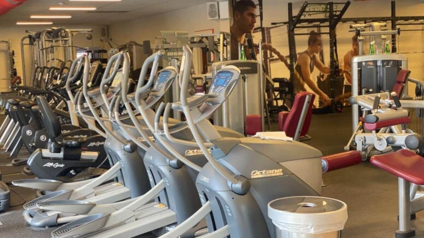 Træningsmaskiner i et moderne fitnesscenter med vægte og spejle i baggrunden.
