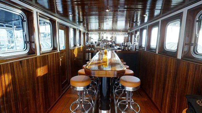 En elegant bar indeni en båd med træpaneler og barstole i læder. Lysindfald fra vinduerne skaber en hyggelig atmosfære.