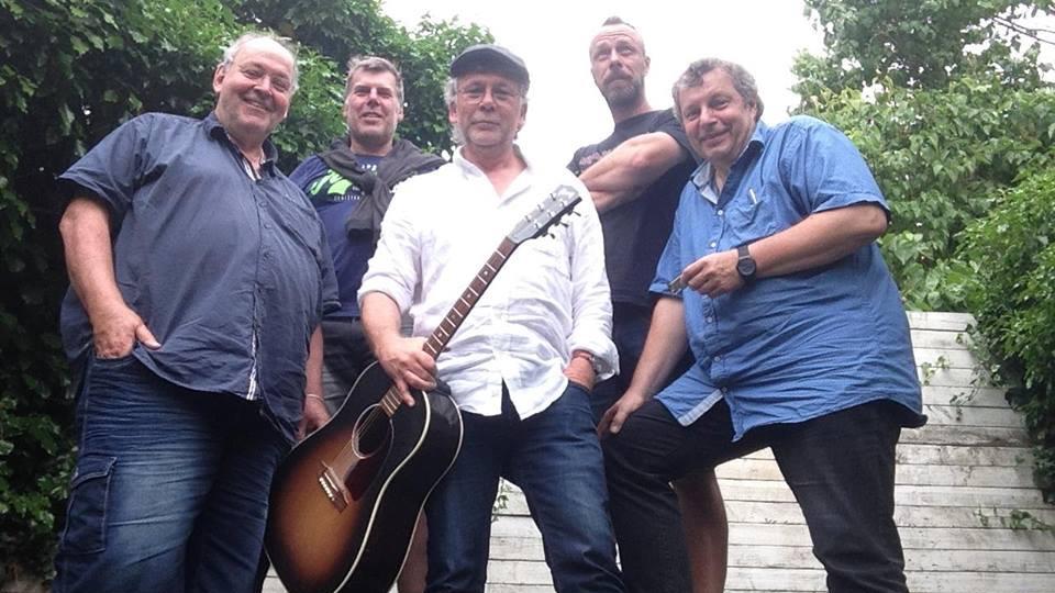Fem mænd står sammen på en trappe, en af dem holder en guitar, alle smiler og ser glade ud.