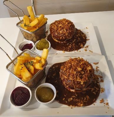 To tallerkener med burgere dækket af sprøde løg og brun sovs, serveret med pomfritter og forskellige saucer.