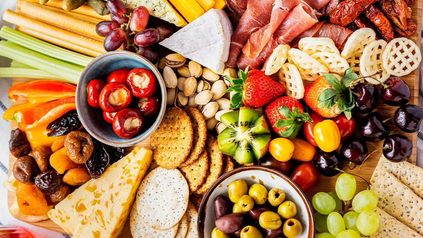 Farverig tapasbræt med frugt, grøntsager, oste, kiks, nødder og skinke i skåle og på et skærebræt.