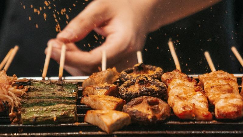 En hånd drysser krydderier over grillspyd med skaldyr, svampe og grøntsager på en grill.
