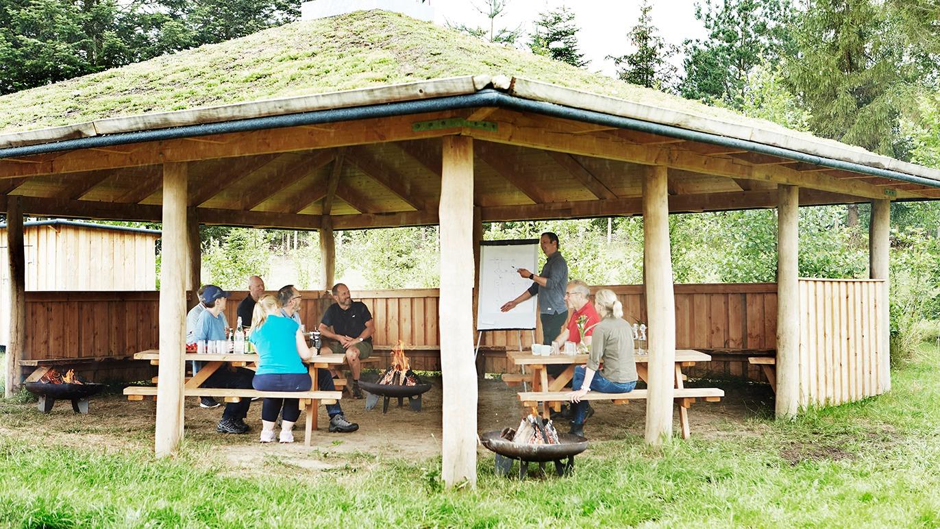 Gruppe mennesker sidder ved borde under en overdækket pavillon i naturen, mens de deltager i en aktivitet.