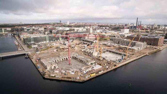 Luftfoto af byggeplads ved vandet med kraner og bygninger under konstruktion i en by.
