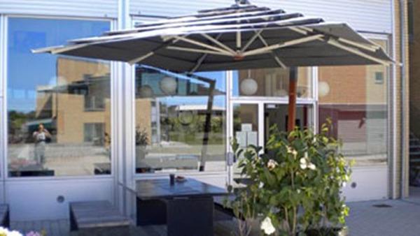 En parasol over et udendørs bord med planter foran en bygning med store vinduer.