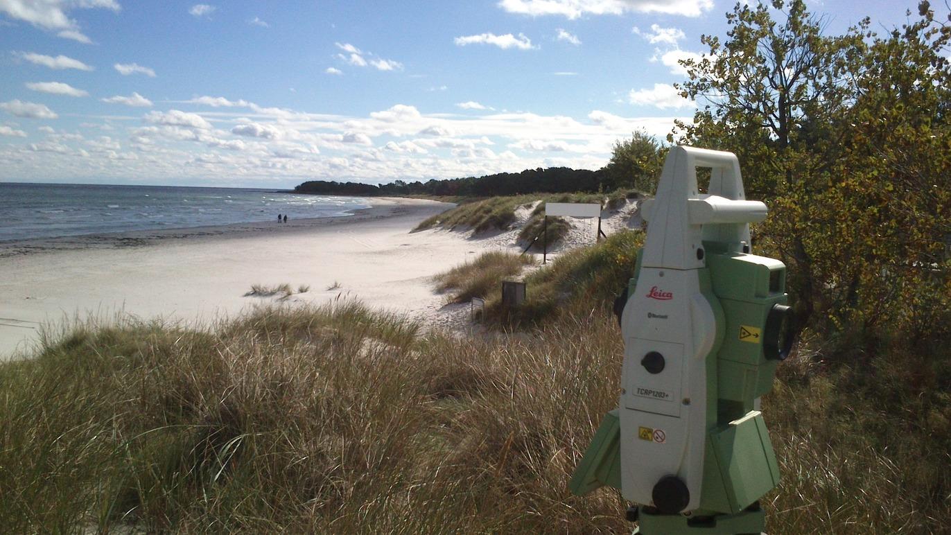 En totalstation står på en sandstrand med bølger og grønne træer i baggrunden under en blå himmel.