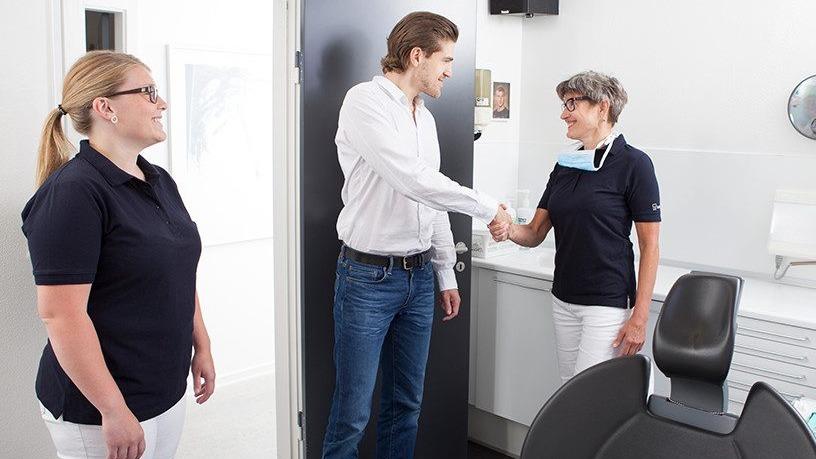 Tre personer i en tandklinik, hvor en mand håndtrykker en kvinde, mens en anden kvinde ser på.