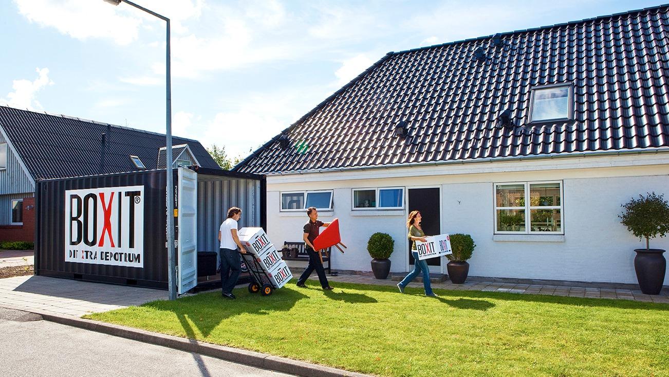 Tre personer bærer kasser ud af en container ved en hvid villa med sort tag i solrigt vejr.