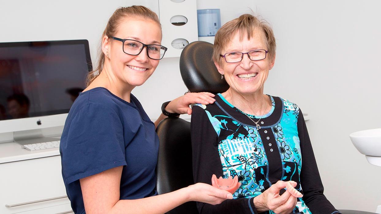 To kvinder smiler til kameraet i en tandlægeklinik, en yngre med briller og en ældre i farverig bluse.