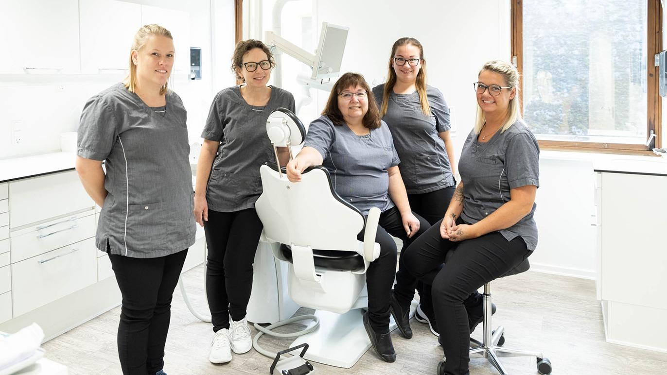 Fem tandlæger står sammen i en tandklinik, iført grå uniformer, med en tandlægestol i fokus.