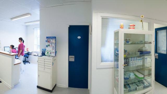 En moderne klinik med en reception og et medicinsk skab fyldt med forsyninger. To personer arbejder ved computeren.