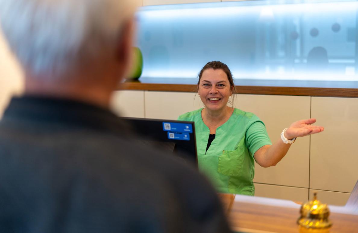 En smilende receptionist i grøn bluse byder velkommen til en gæst ved skranken.