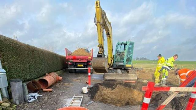 Gravemaskine arbejder på en byggeplads med en lastbil, der leverer sand og arbejdere i sikkerhedstøj.