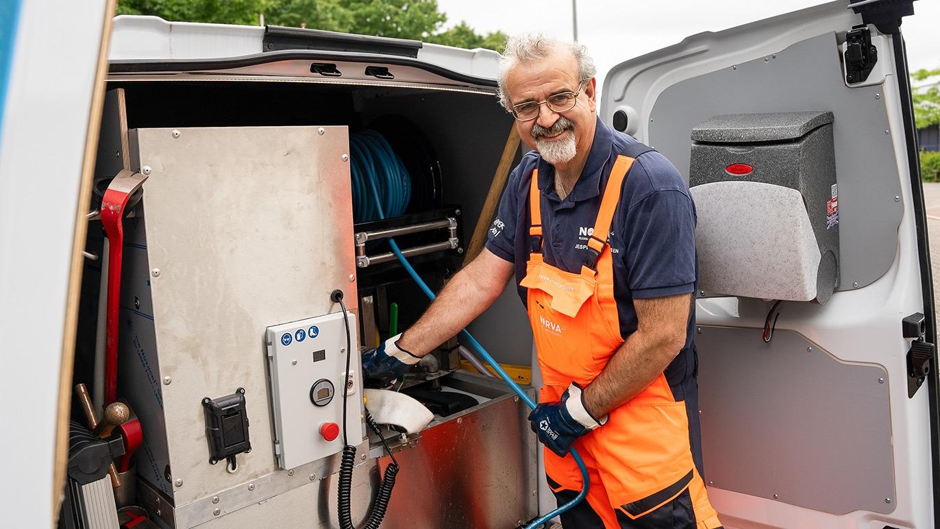 En smilende mand i arbejdstøj står ved en åben varevogn og arbejder med udstyr til rengøring.