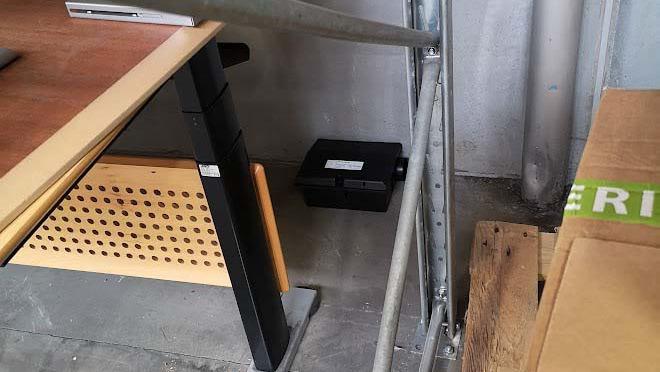 Et hjørne af et rum med et skrivebord, en sort kasse og nogle kasser i baggrunden.