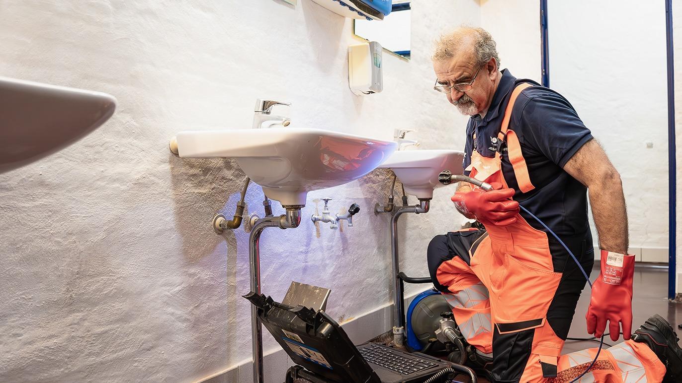 En håndværker i orange arbejdstøj reparerer håndvaske med værktøj i et badeværelse.