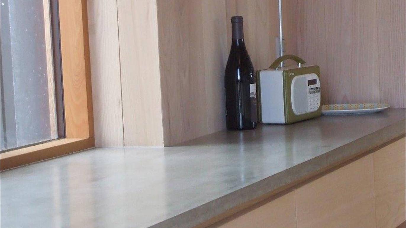 En moderne køkkenbordplade i beton med en vinflaske, en radio og en tallerken ved vinduet.