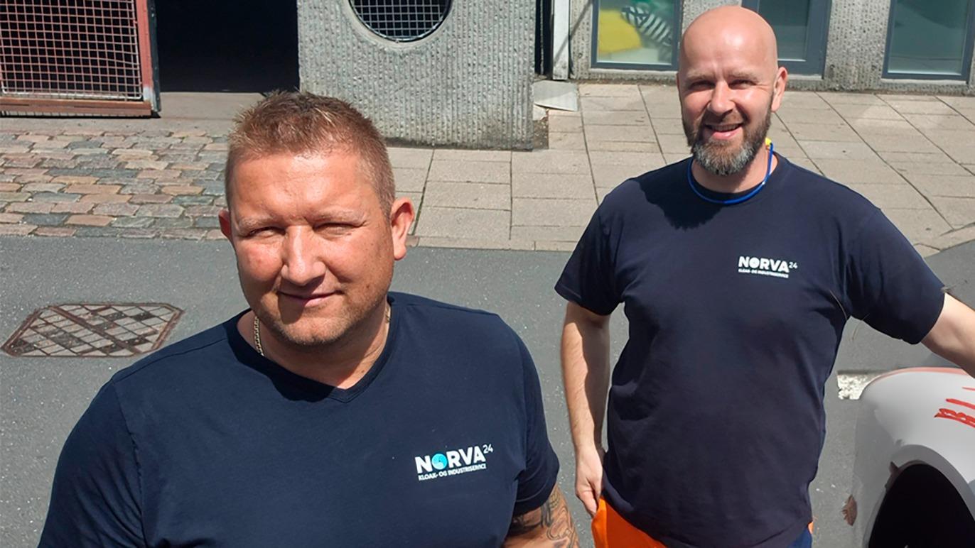 To mænd i mørkeblå t-shirts med logoet NORVA står smilende på en gade med brosten.