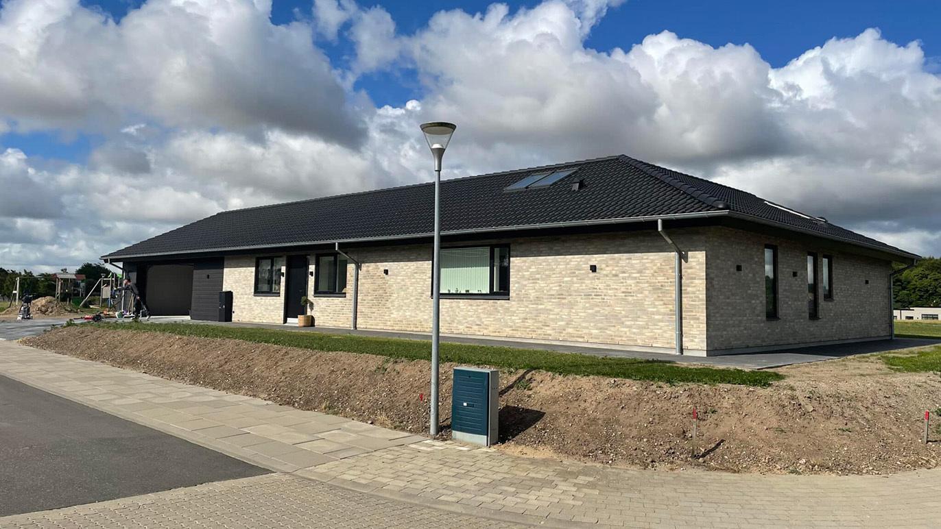 Moderne murstenshus med sort tag og store vinduer, beliggende på en rolig vej under en skyet himmel.